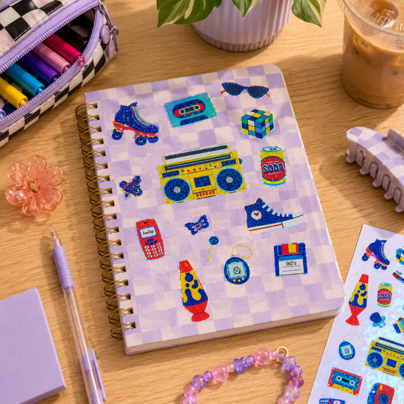 Checkered notebook covered with colourful retro themed holographic stickers including a boom box, lava lamp, vintage mobile phone, cassette tape, roller skate and classic 1990s inspired accessories displayed on a creative workspace.