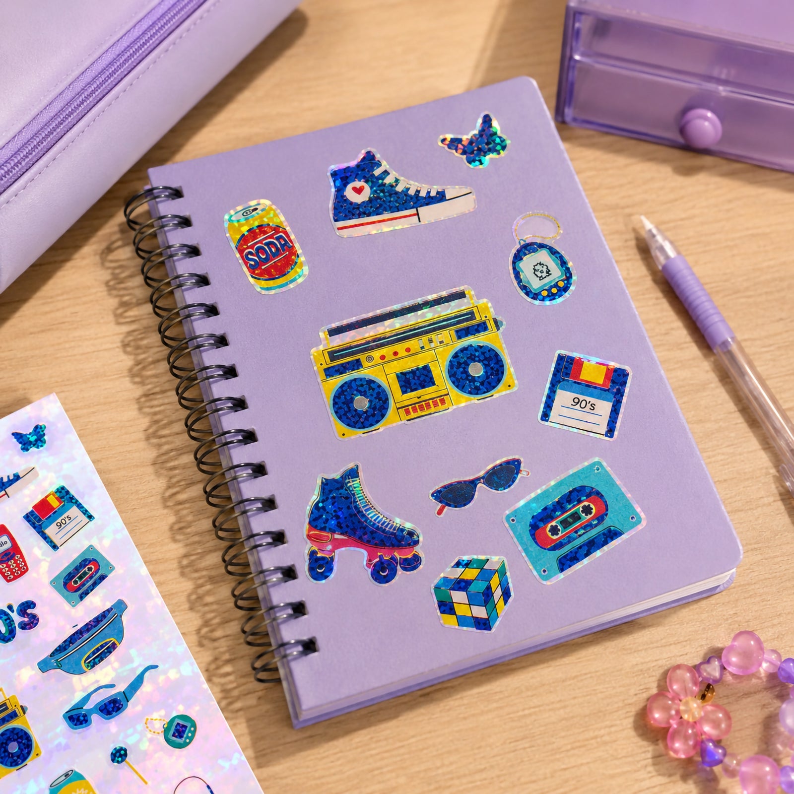 Purple notebook decorated with holographic retro stickers featuring roller skates, a boom box, cassette tape, soda can, floppy disk and sunglasses. The reflective foil finish creates vibrant sparkle against the pastel stationery background.