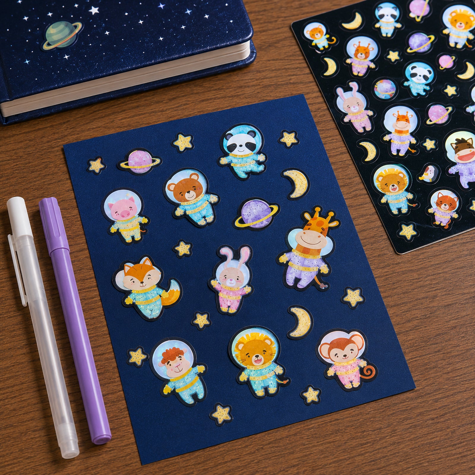 Space themed stickers arranged on dark blue card beside stationery items. Adorable animal astronauts and glowing celestial objects create an engaging scene for creative projects and decorating activities.