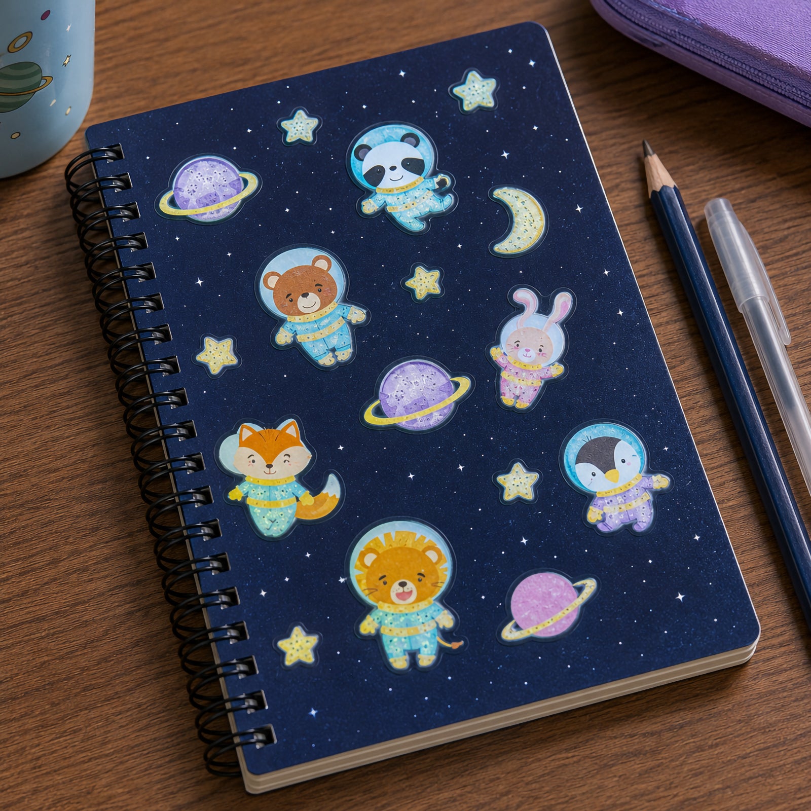 Spiral notebook decorated with glowing space animal stickers including a panda, bear, rabbit, fox, penguin and lion dressed in astronaut suits. Colourful planets, stars and moons create a playful galaxy themed scene.