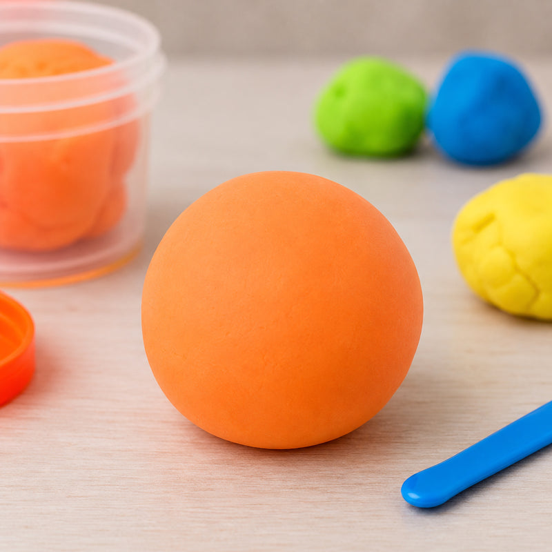 Smooth ball of vibrant fluoro orange CreativeClay displayed on a tabletop beside an open container of clay and sculpting tool. Colourful clay pieces in the background highlight the bright fluorescent appearance and soft texture, showcasing its suitability for creative modelling, classroom crafts and imaginative sculpting projects.