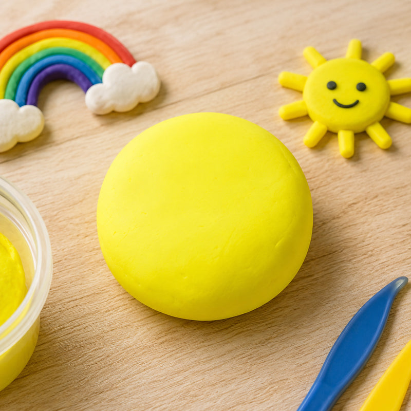 Smooth ball of fluoro yellow CreativeClay displayed on a wooden tabletop beside colourful modelling tools and playful rainbow and sunshine clay decorations. The vibrant fluorescent colour highlights the clay's soft texture and suitability for imaginative craft activities, educational projects and creative modelling.