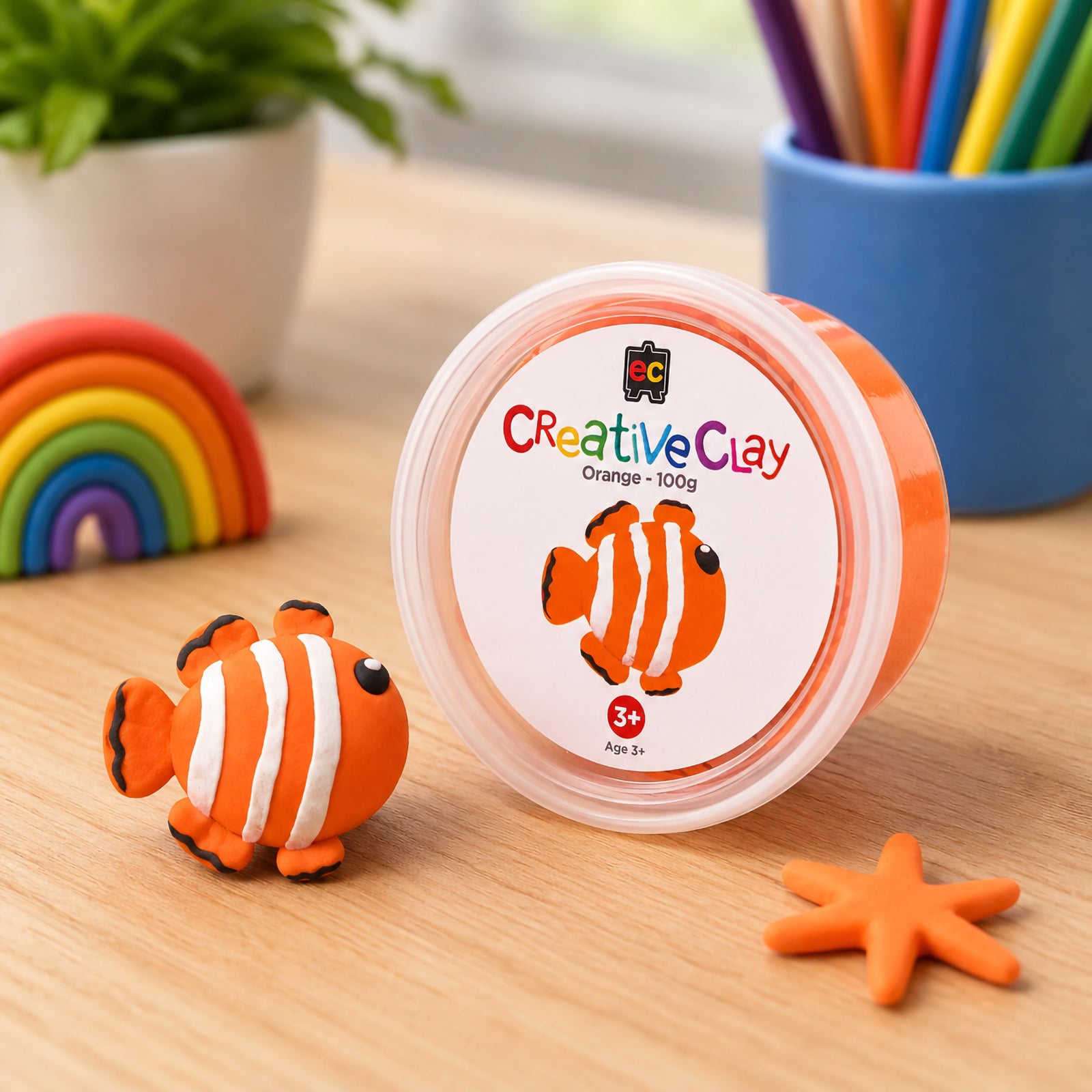 Educational Colours CreativeClay Orange 100g container displayed beside a completed clownfish model, colourful pencils and craft accessories. The image showcases product packaging and creative project inspiration for children and classrooms.