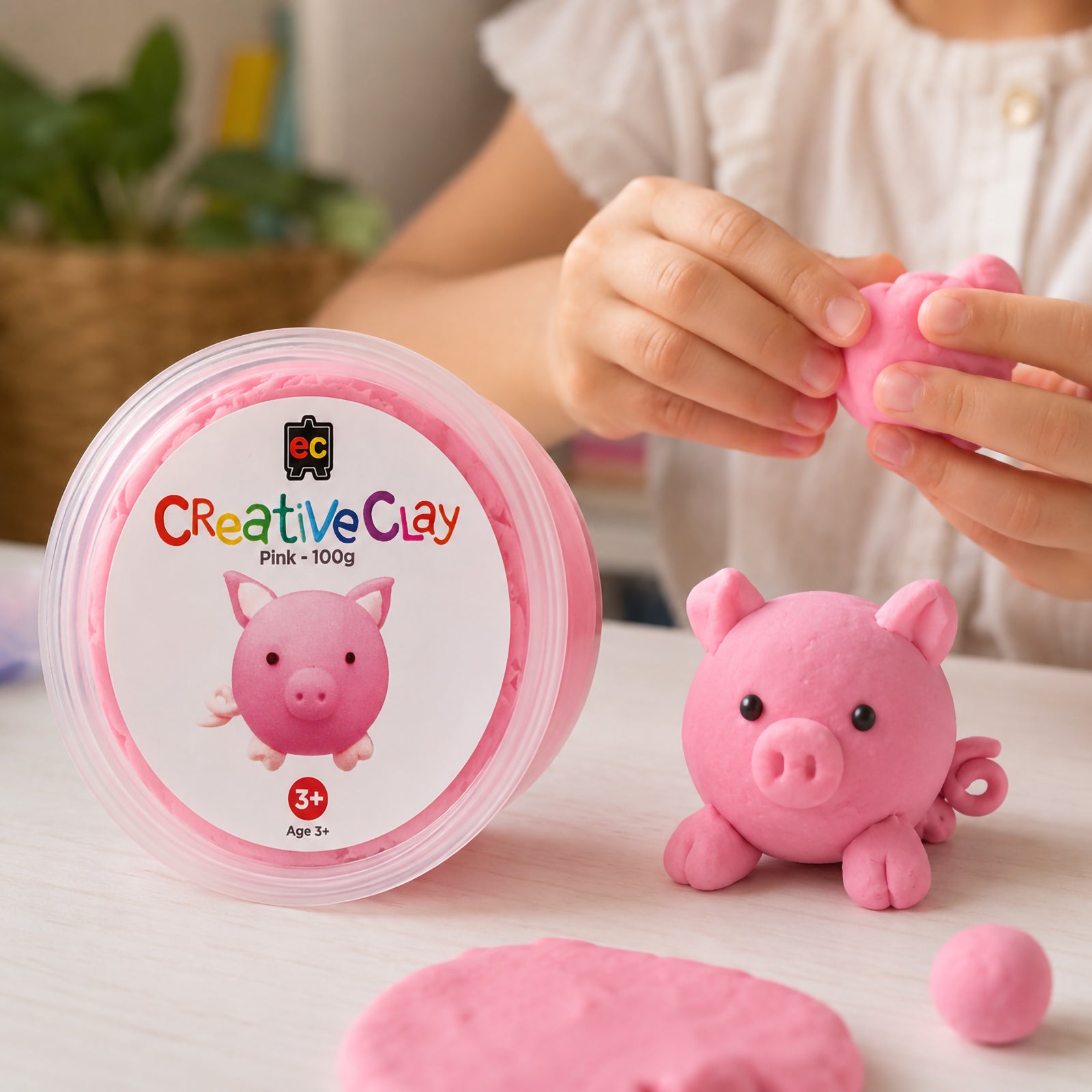Educational Colours CreativeClay Pink 100g container displayed beside a completed pig sculpture while a child shapes additional clay pieces. The image showcases creative play, hands on learning and product packaging.