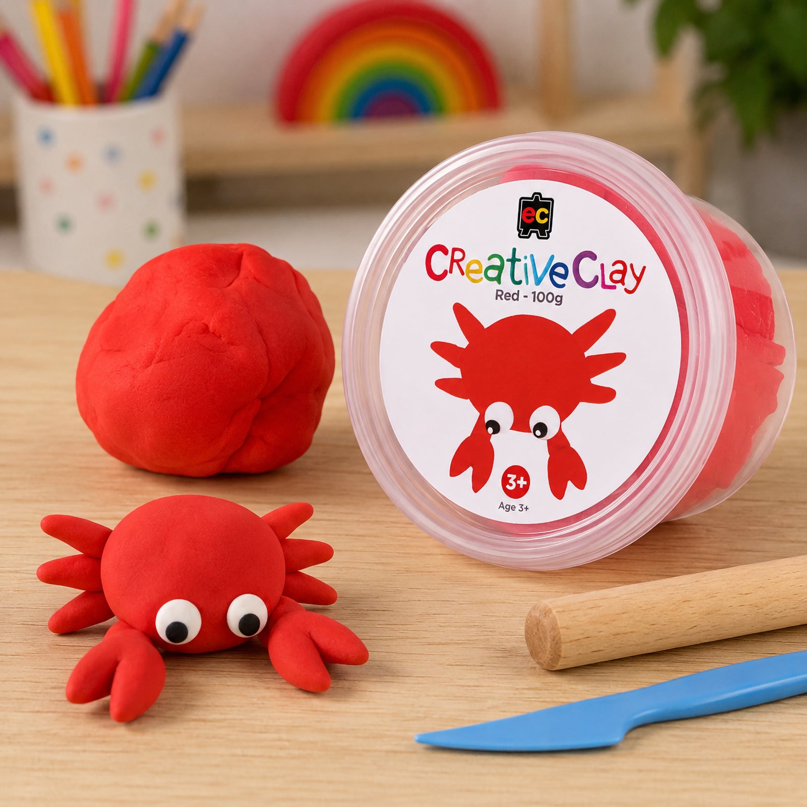 Educational Colours CreativeClay Red 100g container displayed beside completed crab models, modelling tools and craft materials. The scene showcases product packaging and creative inspiration for classroom and home activities.