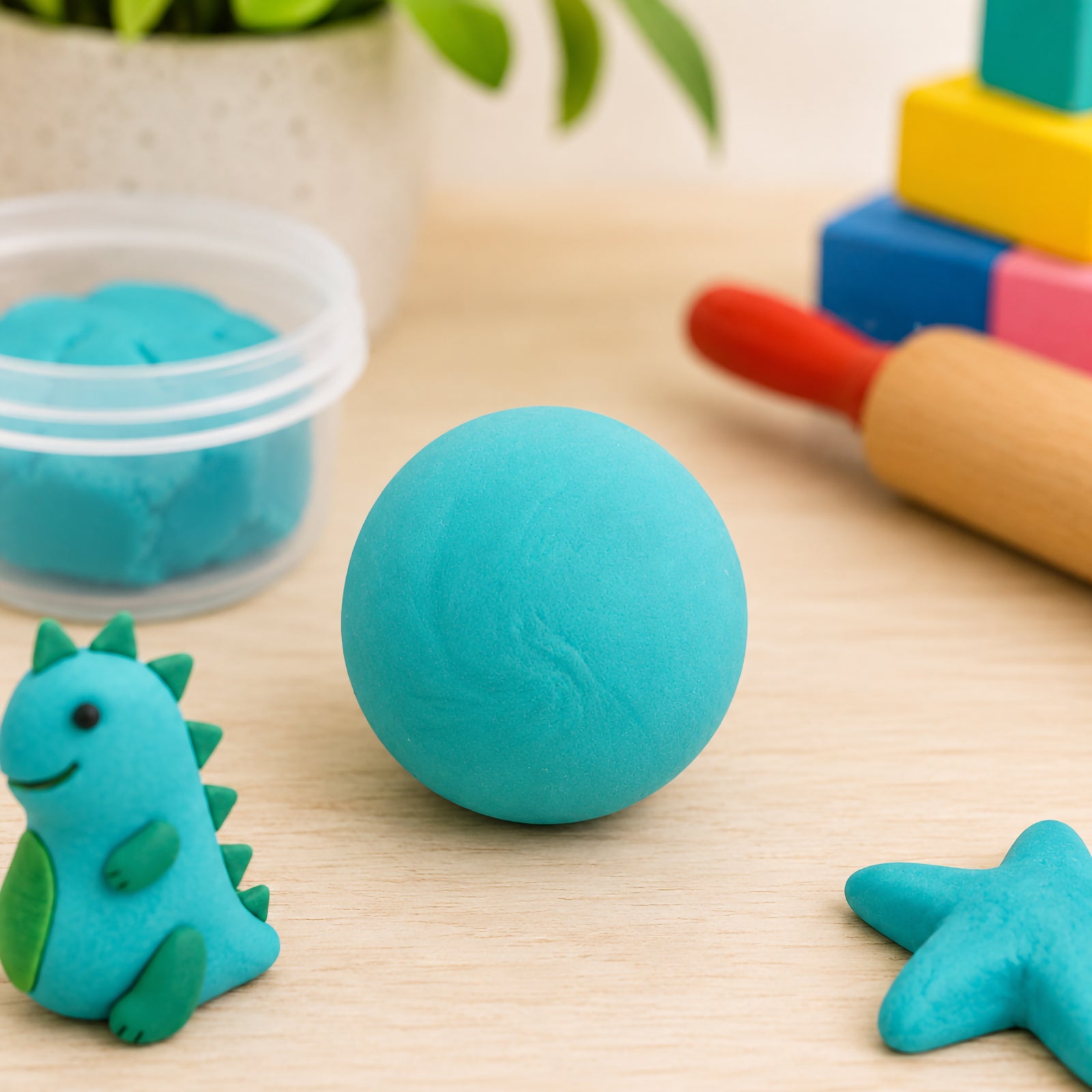 Smooth turquoise CreativeClay rolled into a ball on a craft table beside a small dinosaur model, starfish shape, rolling pin and creative accessories. The image highlights the soft texture, vibrant colour and versatility of the air dry modelling clay for children's art projects.