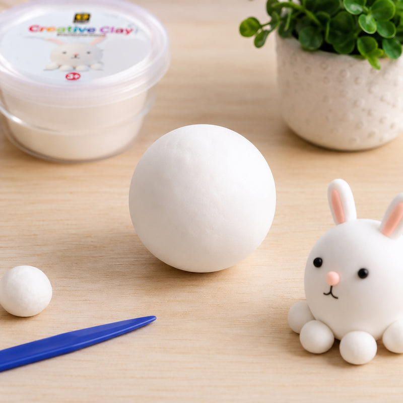 Smooth white CreativeClay rolled into a large ball on a wooden craft table beside a small rabbit model, modelling tool and storage container. The image highlights the soft texture and clean appearance of the air dry clay for creative projects and educational activities.