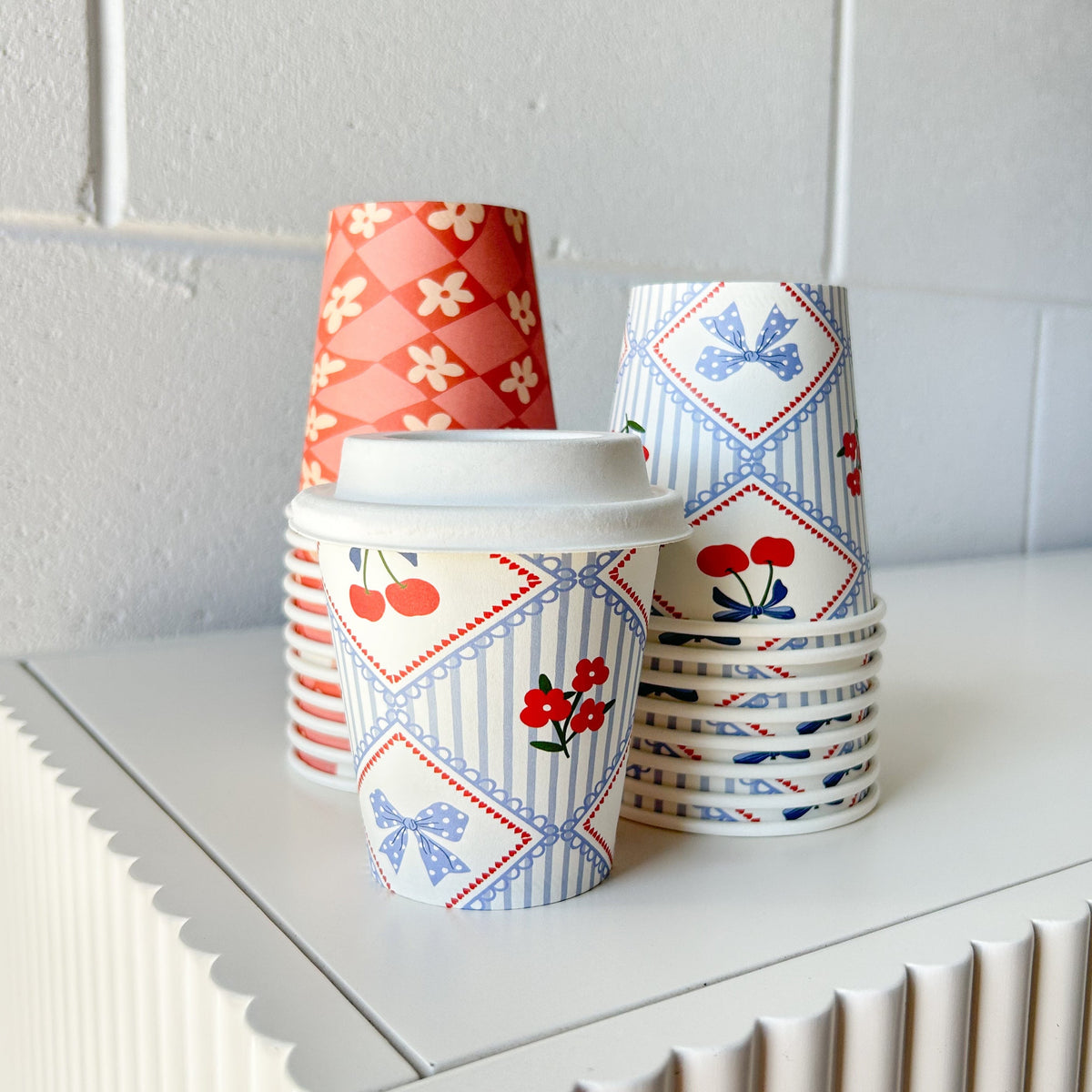 Stacks of Creative Cafe Co 6oz printed takeaway cups with one cup fitted with a white lid on a white cabinet