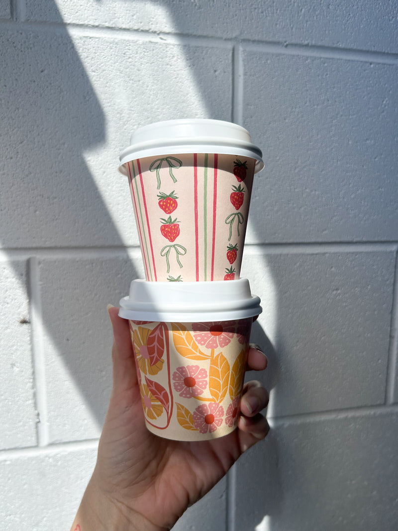 Two Creative Cafe Co 8oz printed takeaway cups with white lids held up in the hand