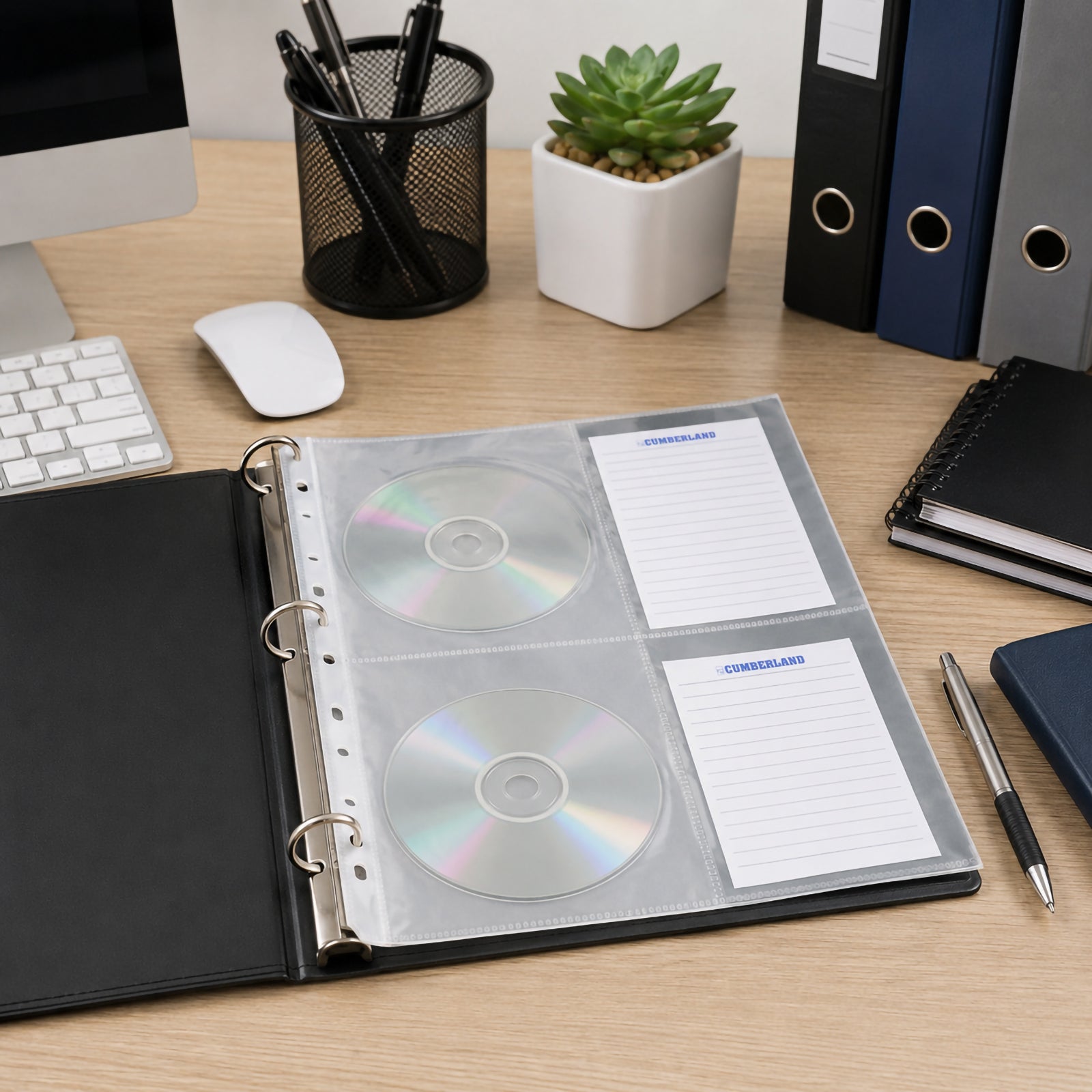 Open ring binder displaying a Cumberland CD DVD storage pocket containing two discs and index cards on a clean office desk with computer, folders, stationery and desk accessories, demonstrating organised media storage.