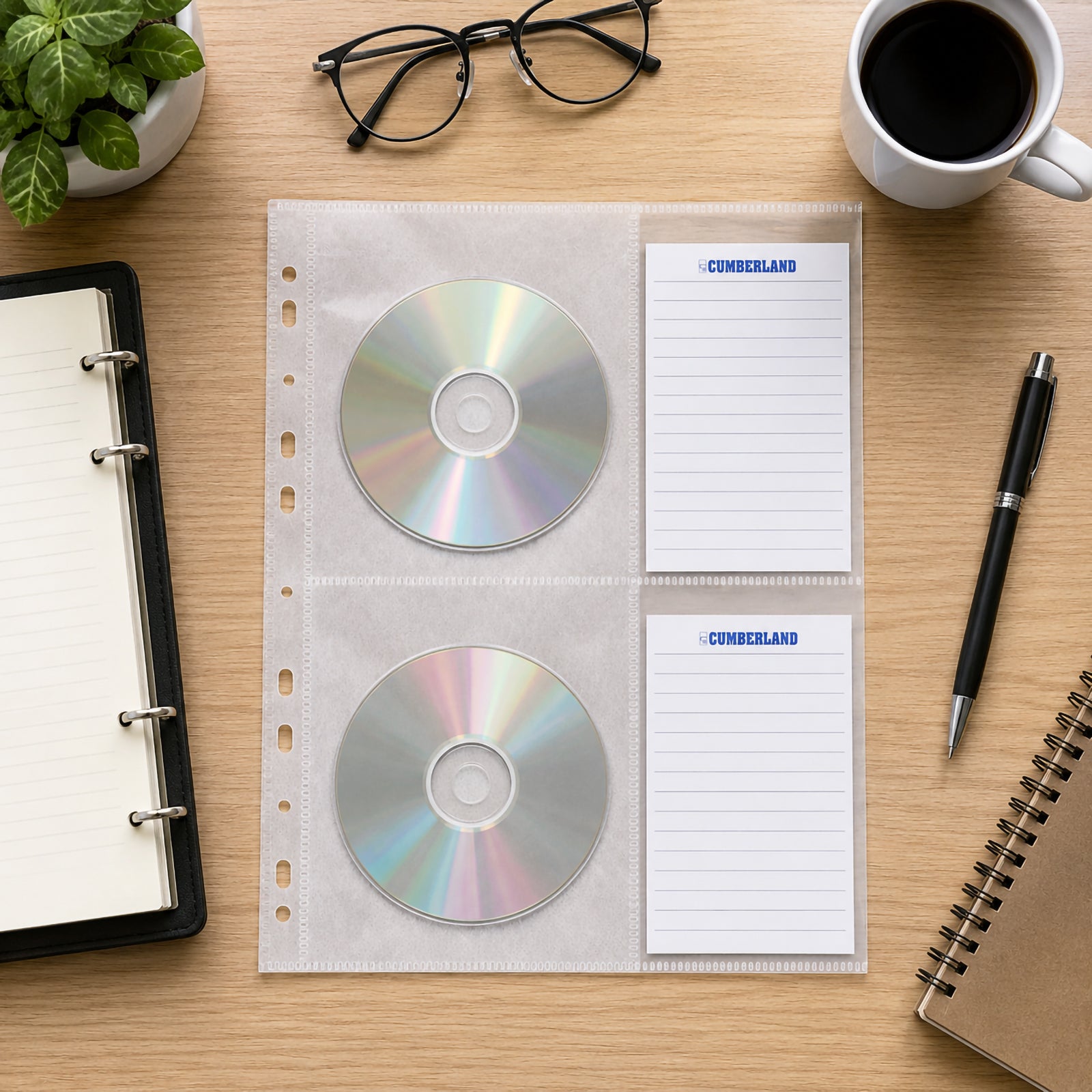Top view of a Cumberland CD DVD binder storage pocket holding two compact discs with matching index cards, positioned on a tidy wooden workspace beside a binder, notebook, coffee cup, glasses and office stationery.