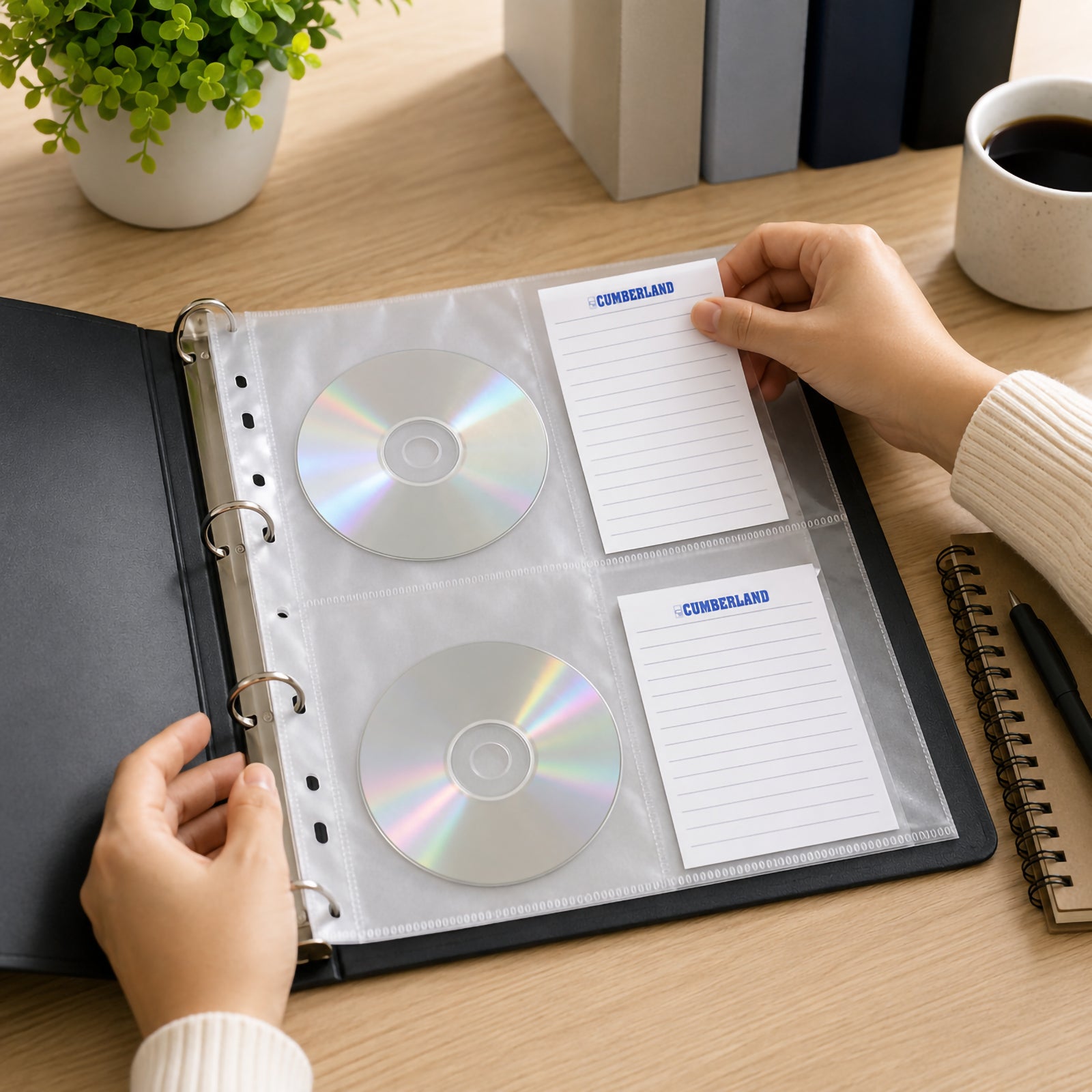 Person organising CDs inside a Cumberland A4 CD DVD binder pocket fitted into a ring binder, with index cards being inserted for easy labelling on a modern office desk alongside stationery, coffee and office accessories.