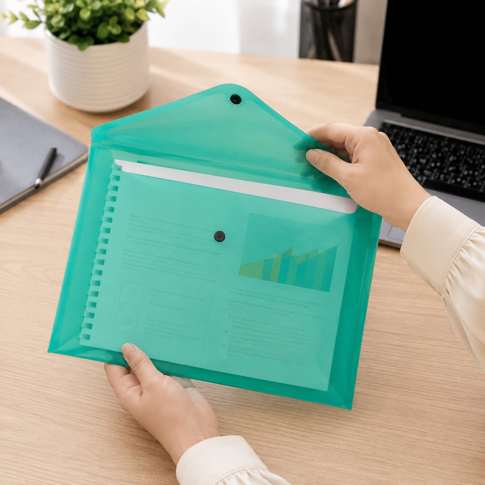 Hands opening a Cumberland A4 transparent green document wallet containing reports and a notebook on a modern office desk beside a laptop, showing the secure press stud closure and generous document capacity.