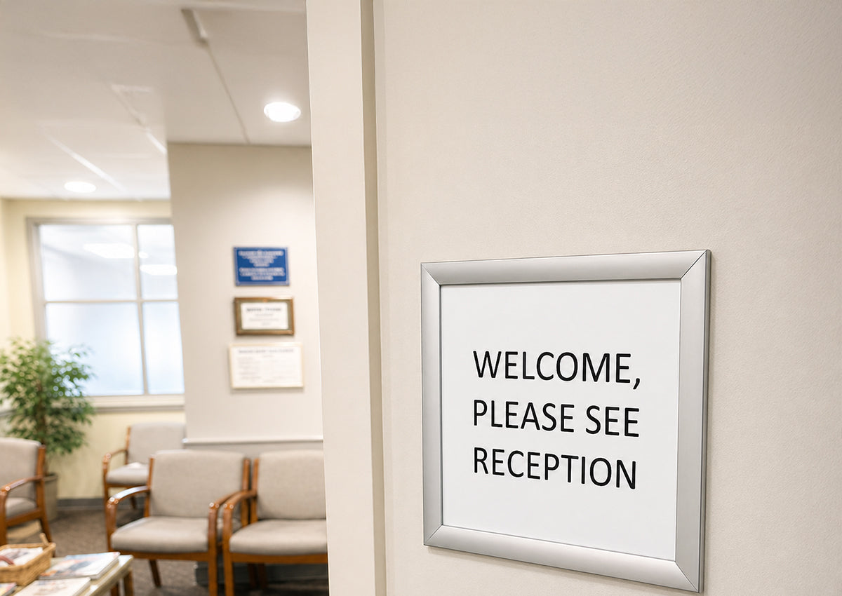 Silver A3 snap frame mounted on a reception wall holding a plain white sign reading Welcome, please see reception, with a waiting area of chairs and a window visible in the background.