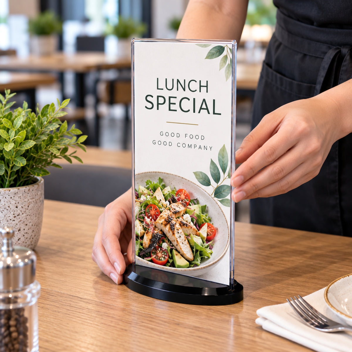 Deflecto DL portrait double sided sign holder being positioned on a timber restaurant table by a hospitality staff member. The clear upright holder contains a lunch special promotion with food imagery and is supported by a compact smoke oval base, demonstrating professional menu presentation in a dining environment.