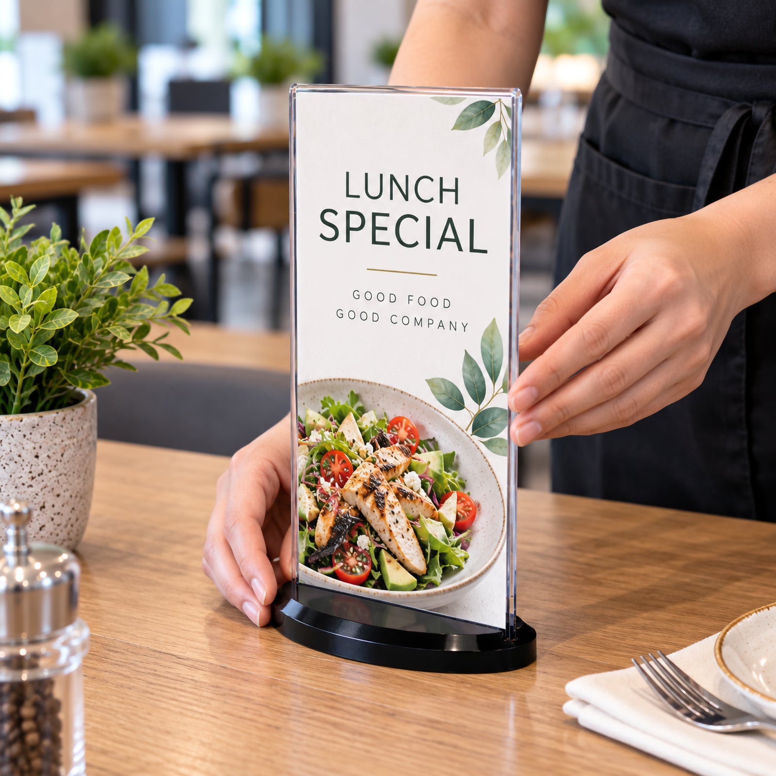 Deflecto DL portrait double sided sign holder being positioned on a timber restaurant table by a hospitality staff member. The clear upright holder contains a lunch special promotion with food imagery and is supported by a compact smoke oval base, demonstrating professional menu presentation in a dining environment.