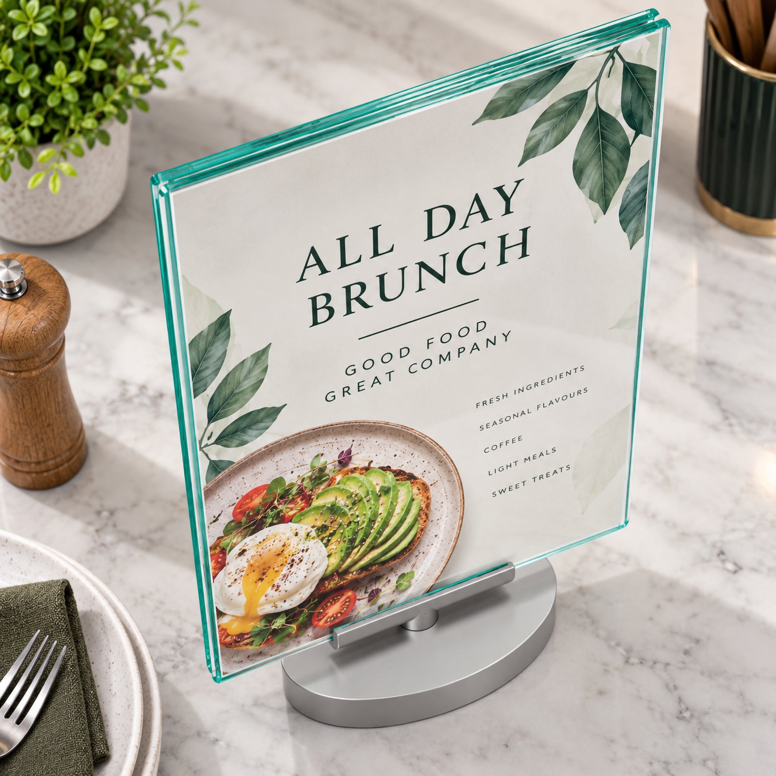 Angled close view of the Deflecto A4 rotating sign holder presenting an all day brunch menu on a marble hospitality counter. The image highlights the polished green tinted edges, clear double sided panel and compact silver base surrounded by tableware and dining accessories.