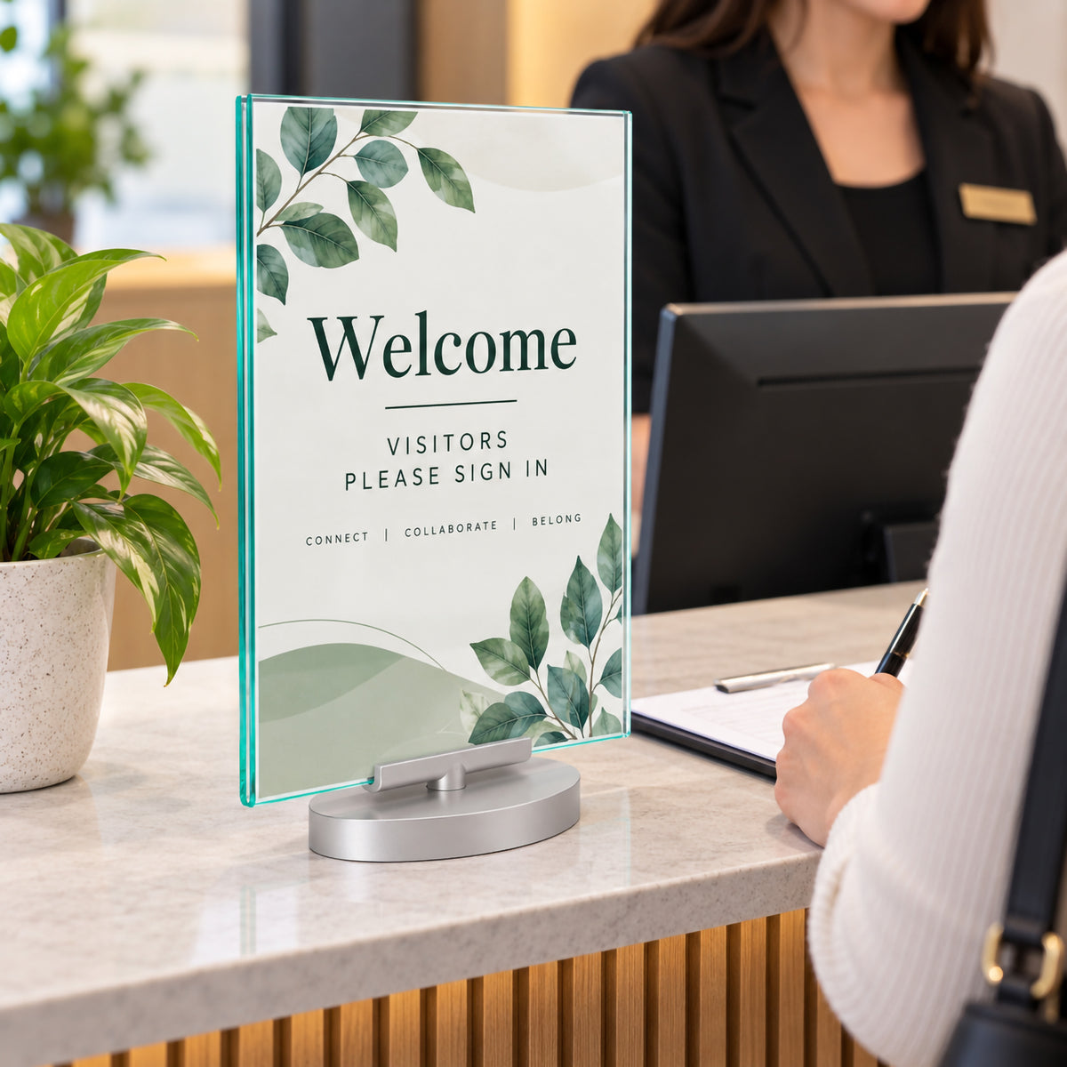 Deflecto A4 portrait double sided rotating sign holder displayed on a modern reception counter with visitor sign in information. The clear glass like panel with subtle green tinted edges stands on a silver rotating base while reception staff assist a visitor in the background.