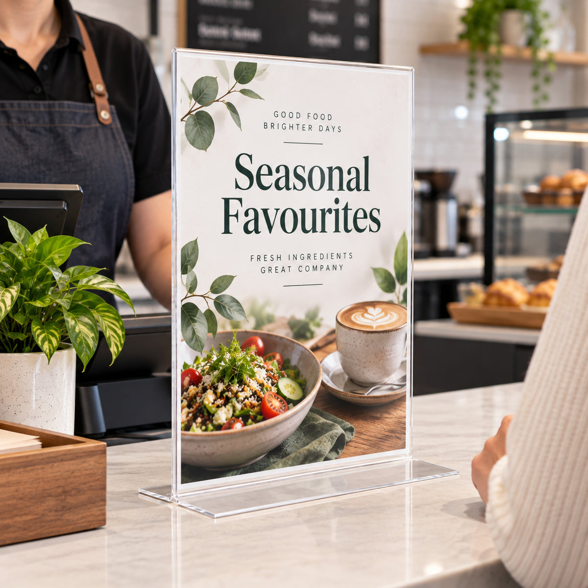 Deflecto A3 portrait double sided T shape sign holder positioned on a cafe service counter displaying seasonal food promotions. The tall clear plastic holder provides prominent customer facing presentation while the wide transparent base keeps the display freestanding beside the point of sale area.