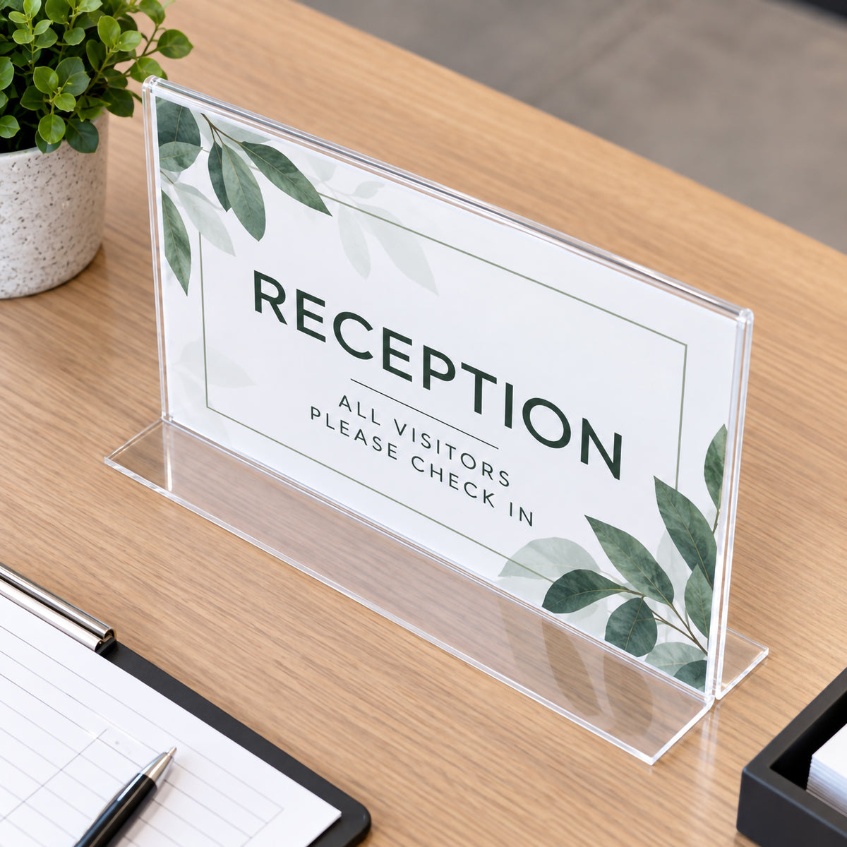 Deflecto A4 landscape clear sign holder positioned on a timber reception desk with visitor check in signage. The angled view highlights the transparent double sided panel and wide flat T shape bottom, showing a professional solution for reception instructions, office notices and customer information.