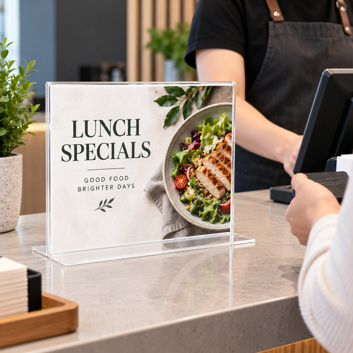 Deflecto A4 landscape double sided T shape sign holder displayed on a cafe counter with a lunch specials insert featuring fresh food imagery. The clear freestanding holder sits beside the point of sale area while a staff member serves a customer, demonstrating practical use for menus and daily promotions.
