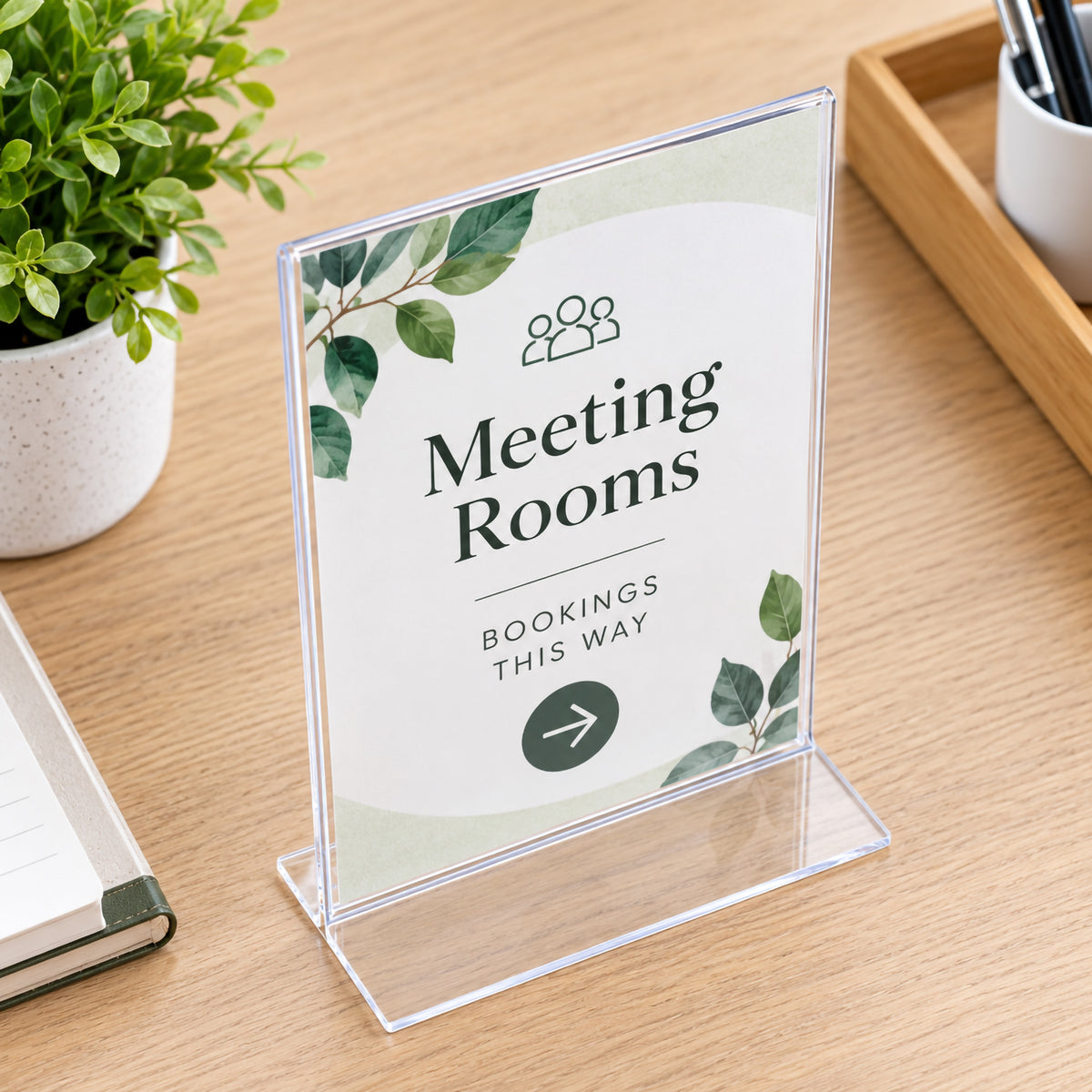 Deflecto A5 portrait clear sign holder viewed from an elevated angle on a timber office desk. The insert directs visitors towards meeting rooms, while nearby stationery and greenery create a professional workplace setting. The transparent edges and broad flat T shape bottom are clearly visible.