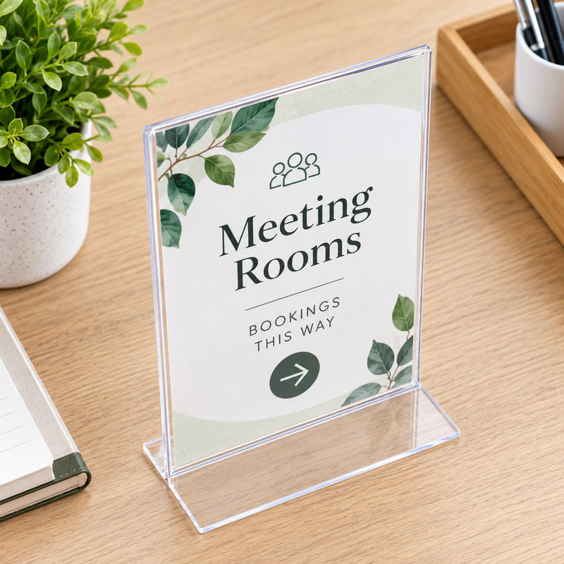 Deflecto A5 portrait clear sign holder viewed from an elevated angle on a timber office desk. The insert directs visitors towards meeting rooms, while nearby stationery and greenery create a professional workplace setting. The transparent edges and broad flat T shape bottom are clearly visible.