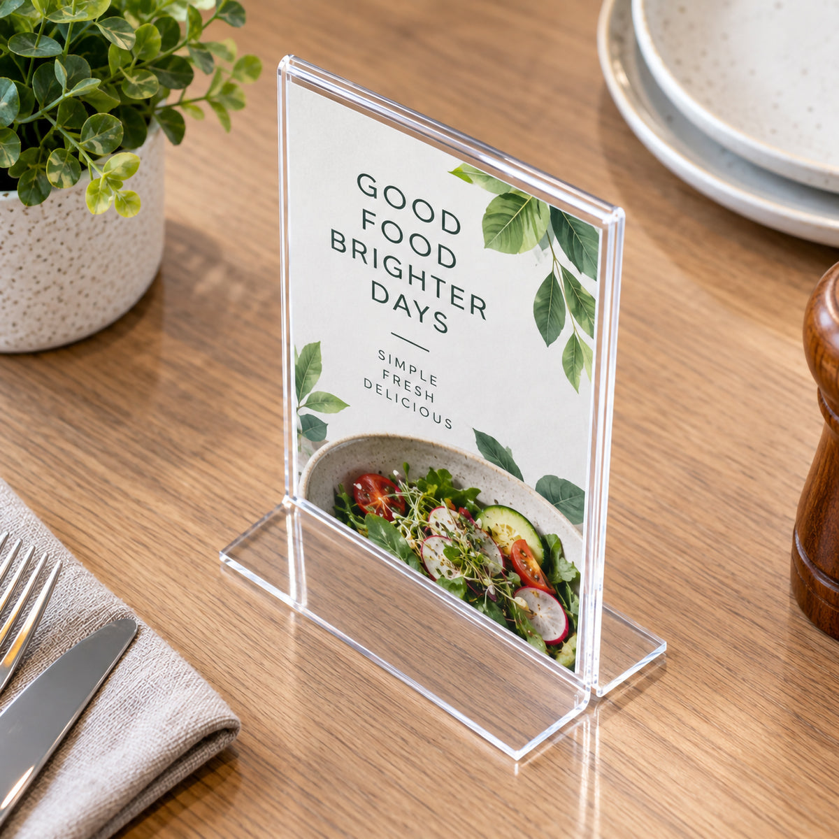 Deflecto clear A6 portrait sign holder viewed from an elevated angle on a timber dining table. The transparent T shape holder presents a fresh food message with salad imagery and is surrounded by tableware, greenery and dining accessories, showing its compact footprint in a restaurant setting.