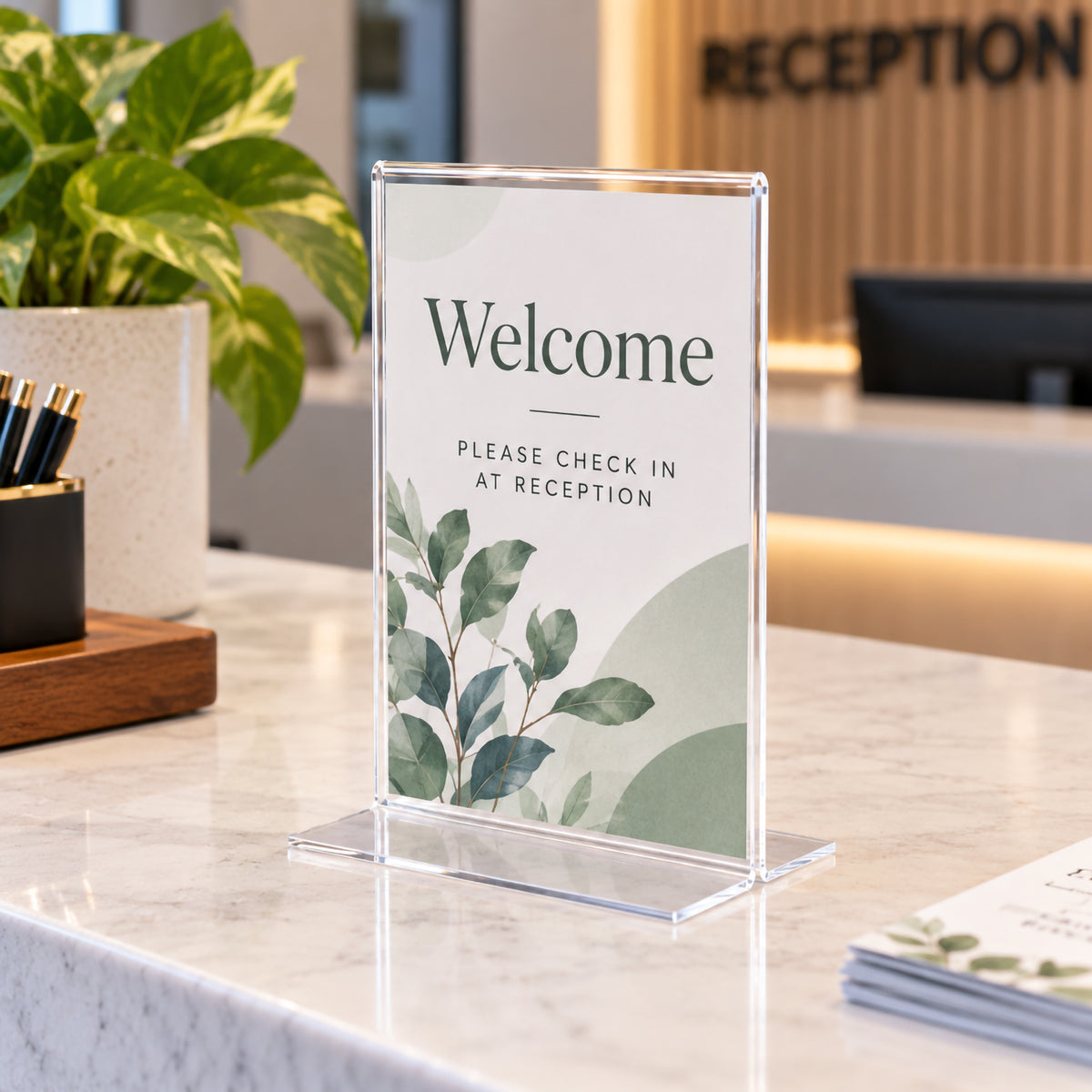 Deflecto A6 portrait T shape sign holder positioned on a modern reception counter. The clear freestanding display contains a welcome and check in message with botanical artwork, demonstrating a neat way to communicate visitor instructions while complementing a professional reception environment.
