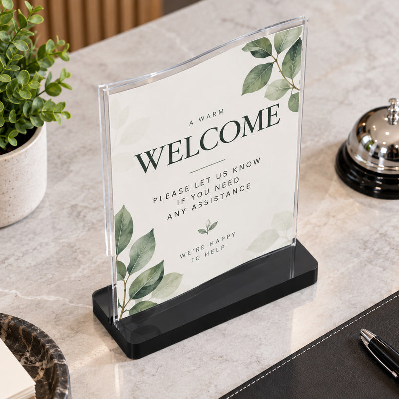 Close angled view of the Deflecto A5 portrait wavy sign holder positioned on a stone reception counter. The clear double sided holder contains a botanical welcome notice and sits securely in its black rectangular base beside reception accessories, showing the distinctive curved upper profile.