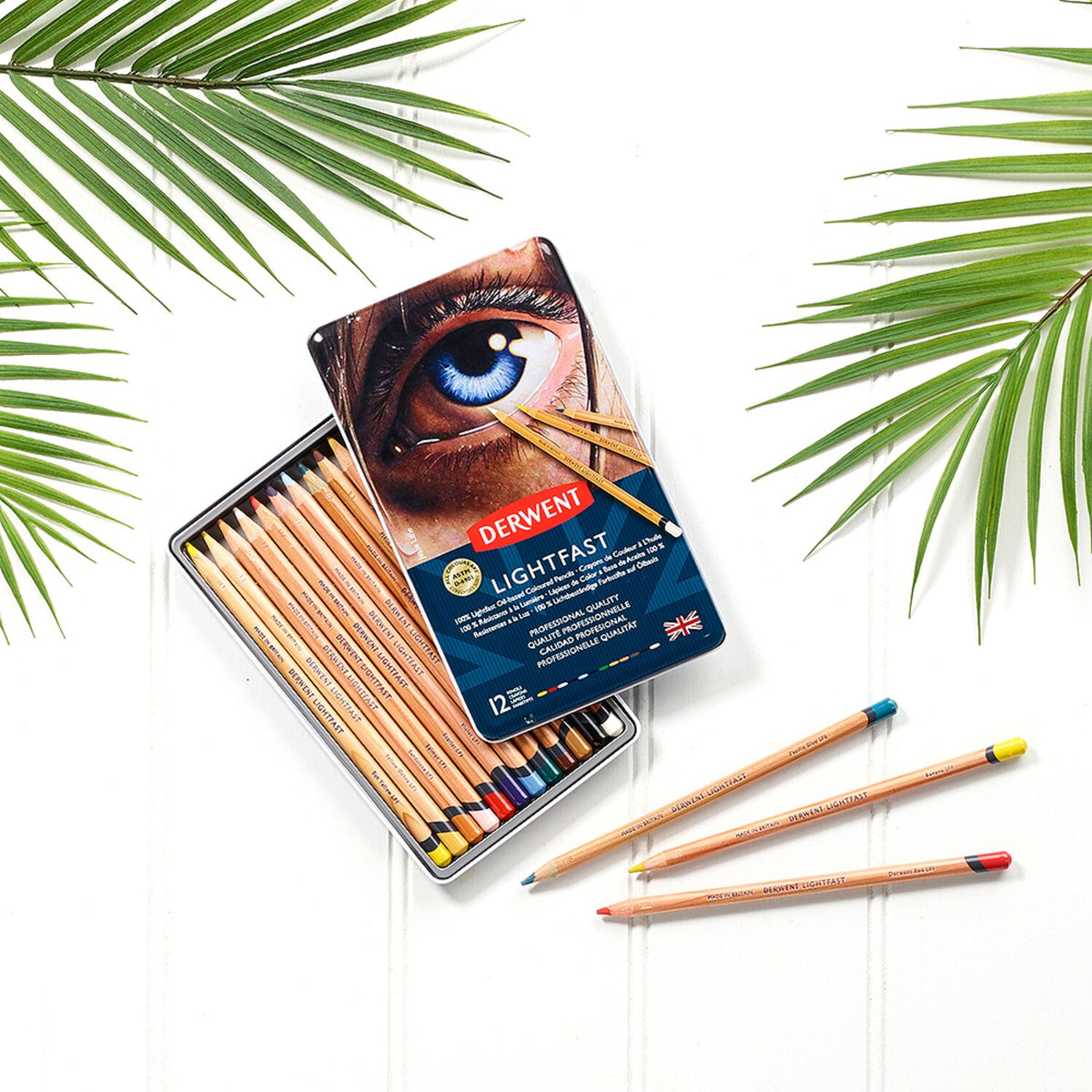 Derwent Lightfast 12 tin open on a white surface with palm leaves, the lid resting on the pencil tray and three loose pencils in front