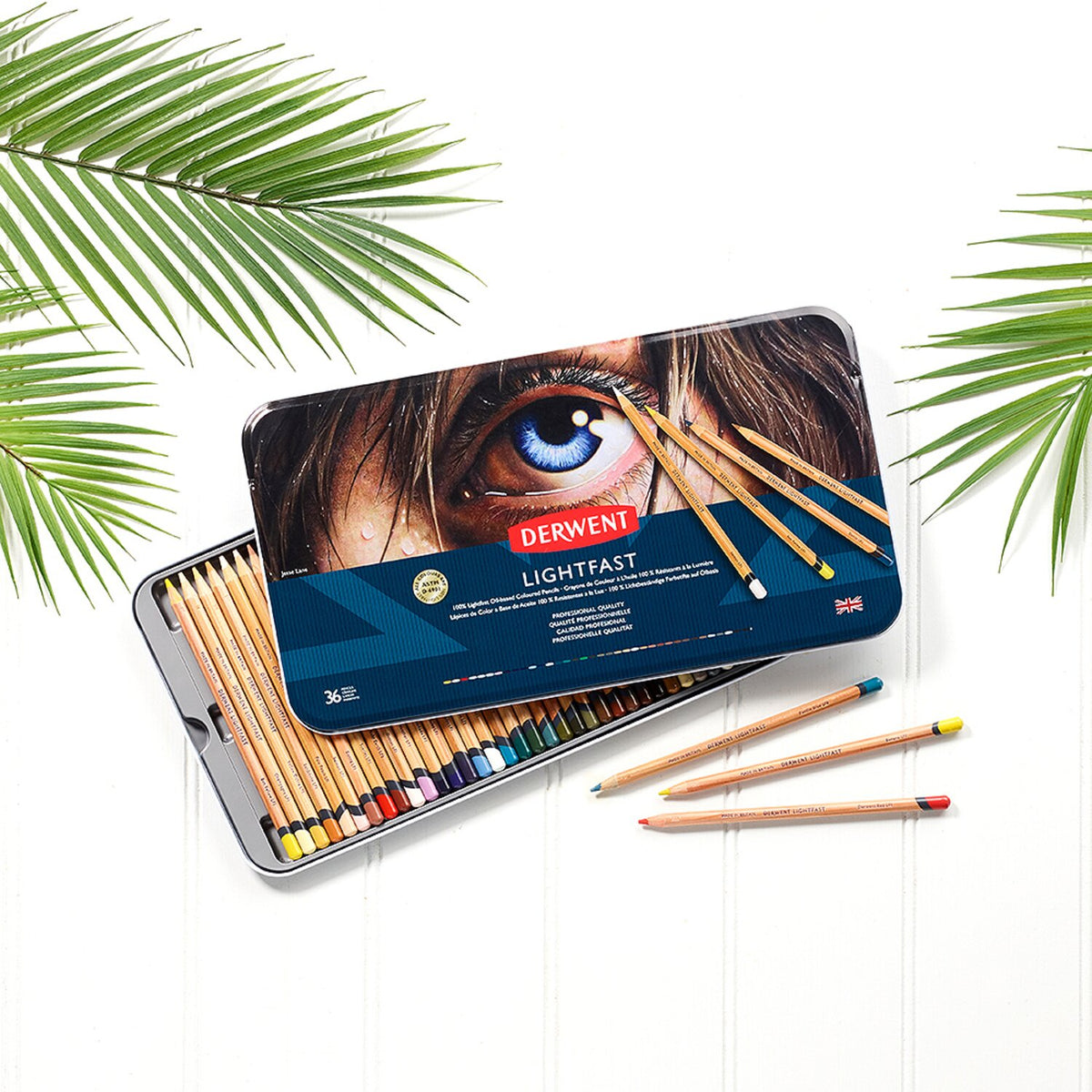Derwent Lightfast 36 tin open on a white surface with palm leaves, the lid resting across the pencil tray and loose pencils in front
