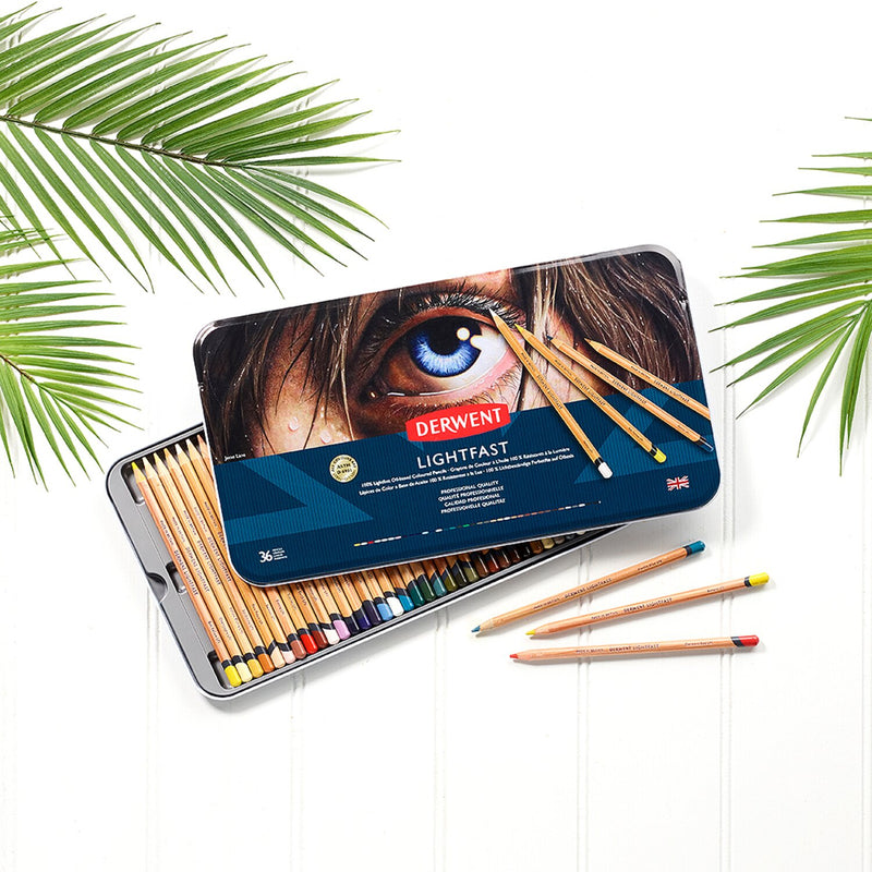 Derwent Lightfast 36 tin open on a white surface with palm leaves, the lid resting across the pencil tray and loose pencils in front