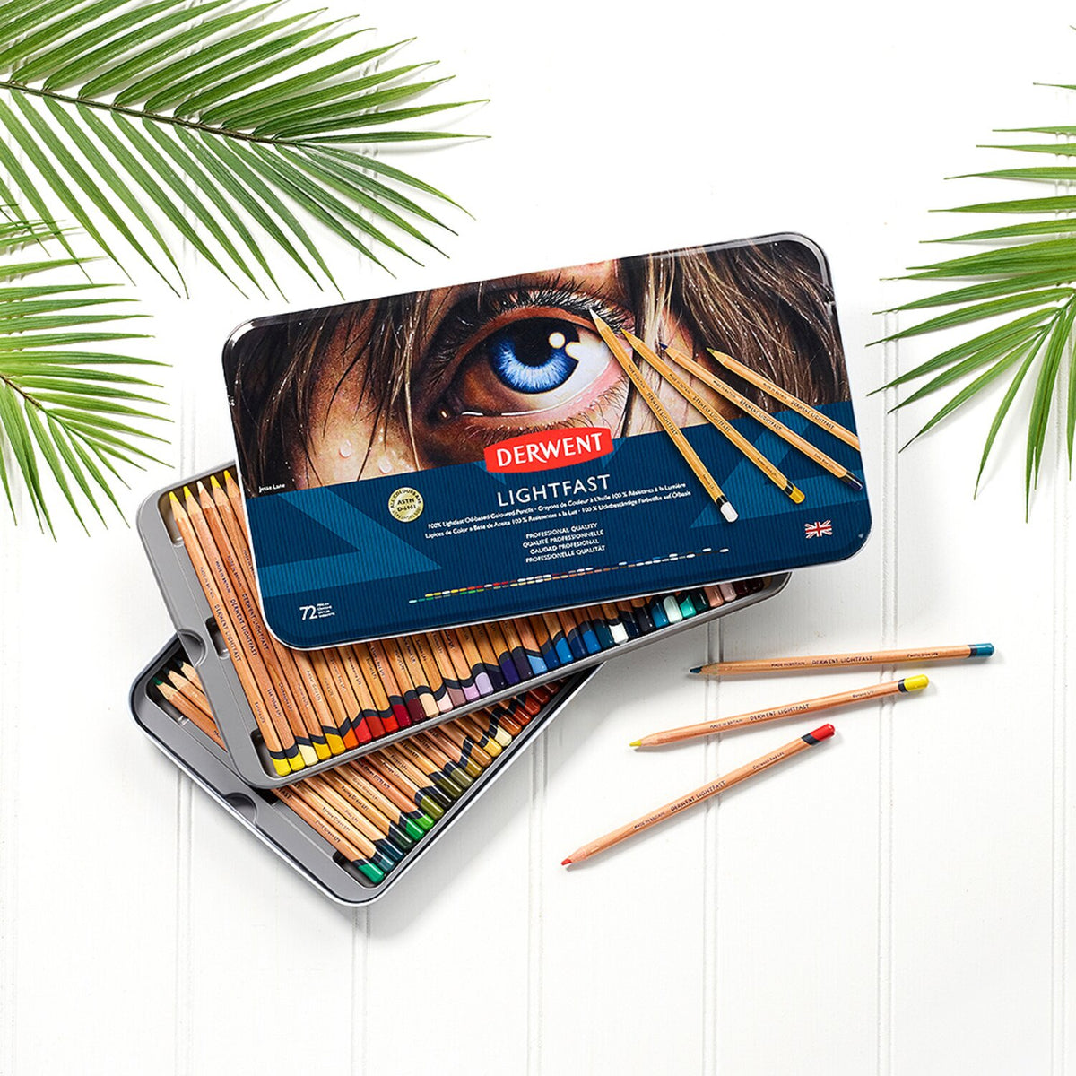 Derwent Lightfast 72 tin open on a white surface with palm leaves, the lid on top of the stacked trays and loose pencils in front