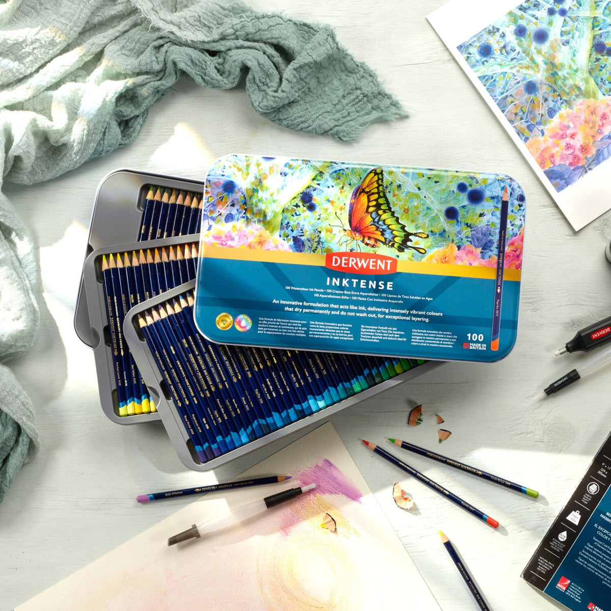 Derwent Inktense 100 pencil tin open on a table beside a butterfly painting, loose Inktense pencils and pencil shavings