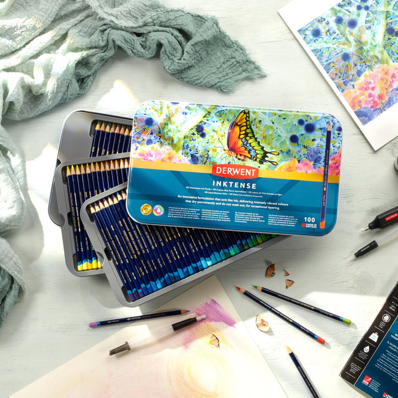 Derwent Inktense 100 pencil tin open on a table beside a butterfly painting, loose Inktense pencils and pencil shavings