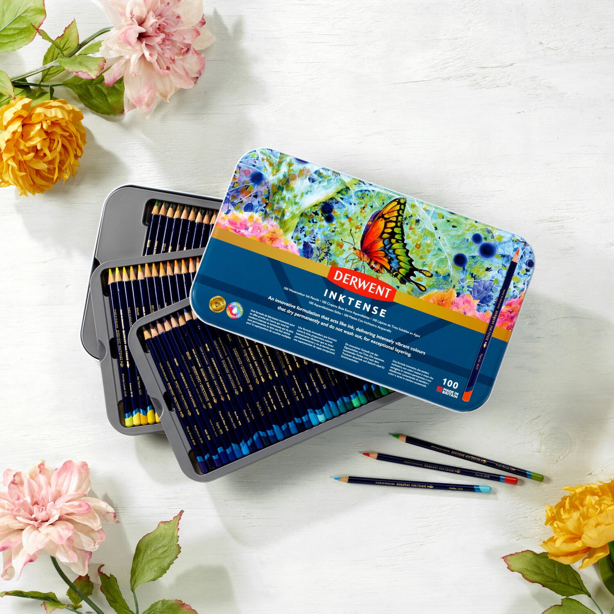 Derwent Inktense 100 pencil tin with its butterfly lid over three trays of pencils, set among flowers with three loose pencils