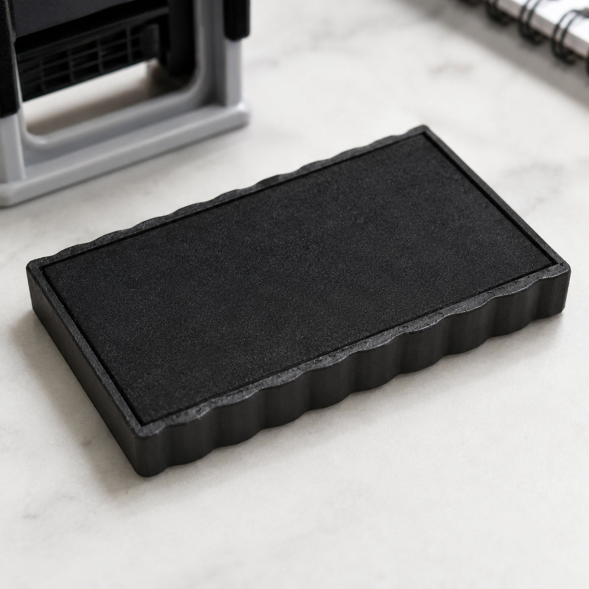 Close up view of the Deskmate Replacement Ink Pad 5 Line 3mm 4mm Text Black resting on a clean desktop beside a self inking stamp. The detailed textured ink surface highlights the genuine replacement pad designed to restore sharp and consistent stamp impressions.