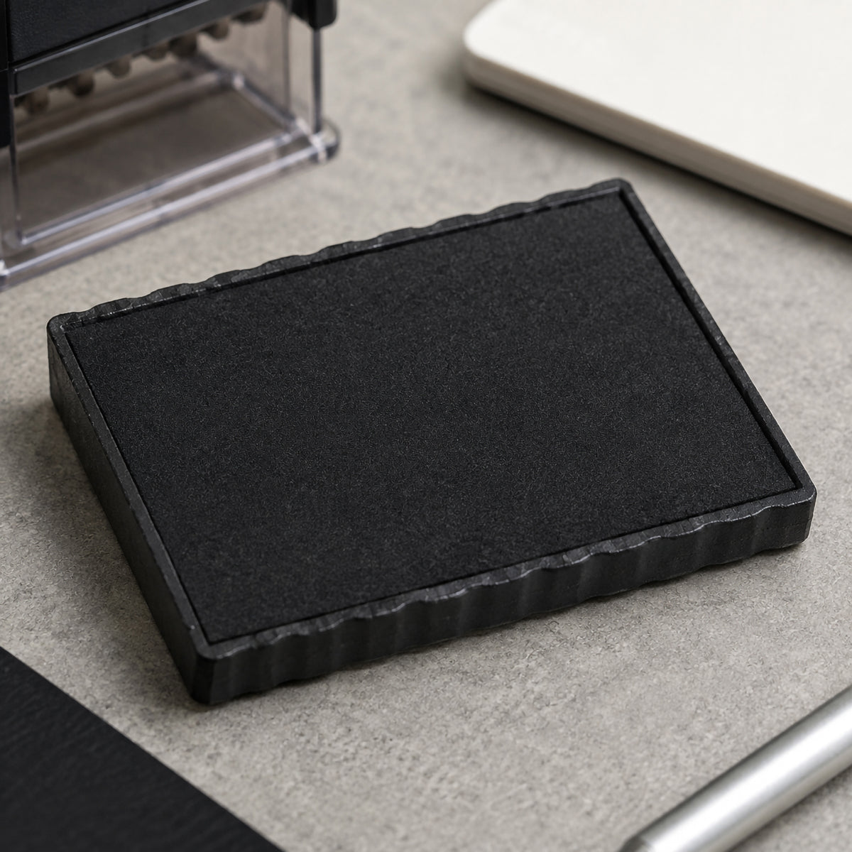 Close up view of the Deskmate Replacement Ink Pad 6 Line 3mm 4mm Text Black resting on a modern office desk beside a compatible self inking stamp. The textured ink surface and durable construction demonstrate the genuine replacement pad designed for long lasting, professional stamp impressions.
