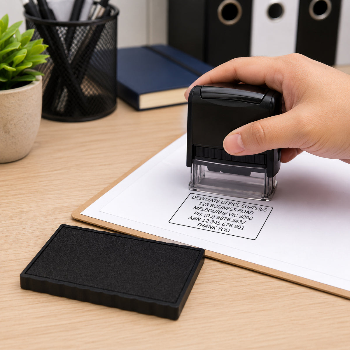 A lifestyle office scene showing a hand using a Deskmate self inking stamp fitted with the Deskmate Replacement Ink Pad 6 Line 3mm 4mm Text Black to create a crisp business address impression on a document. The replacement ink pad sits beside the stamp, highlighting quick replacement and reliable everyday office performance.