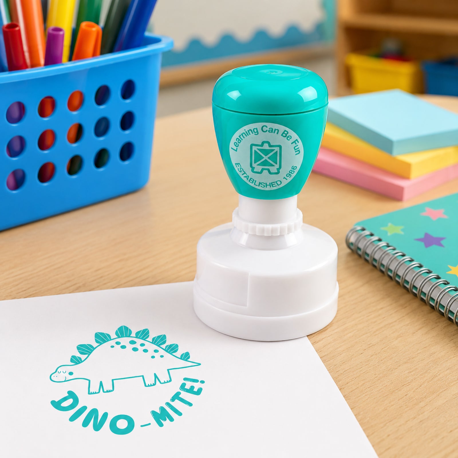 Self inking dinosaur merit stamp shown in an educational setting with coloured markers, notebooks and learning resources. The bright teal impression provides positive reinforcement for effort, participation and progress.