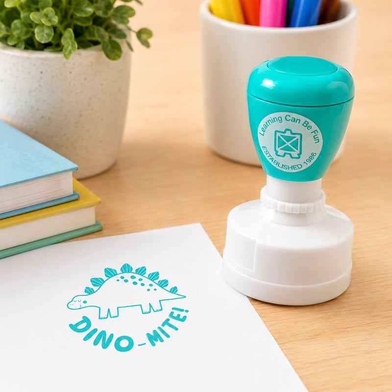 Dino Mite reward stamp positioned near classroom supplies with a vibrant dinosaur design stamped onto white paper. The playful prehistoric theme helps motivate students and celebrate learning achievements.