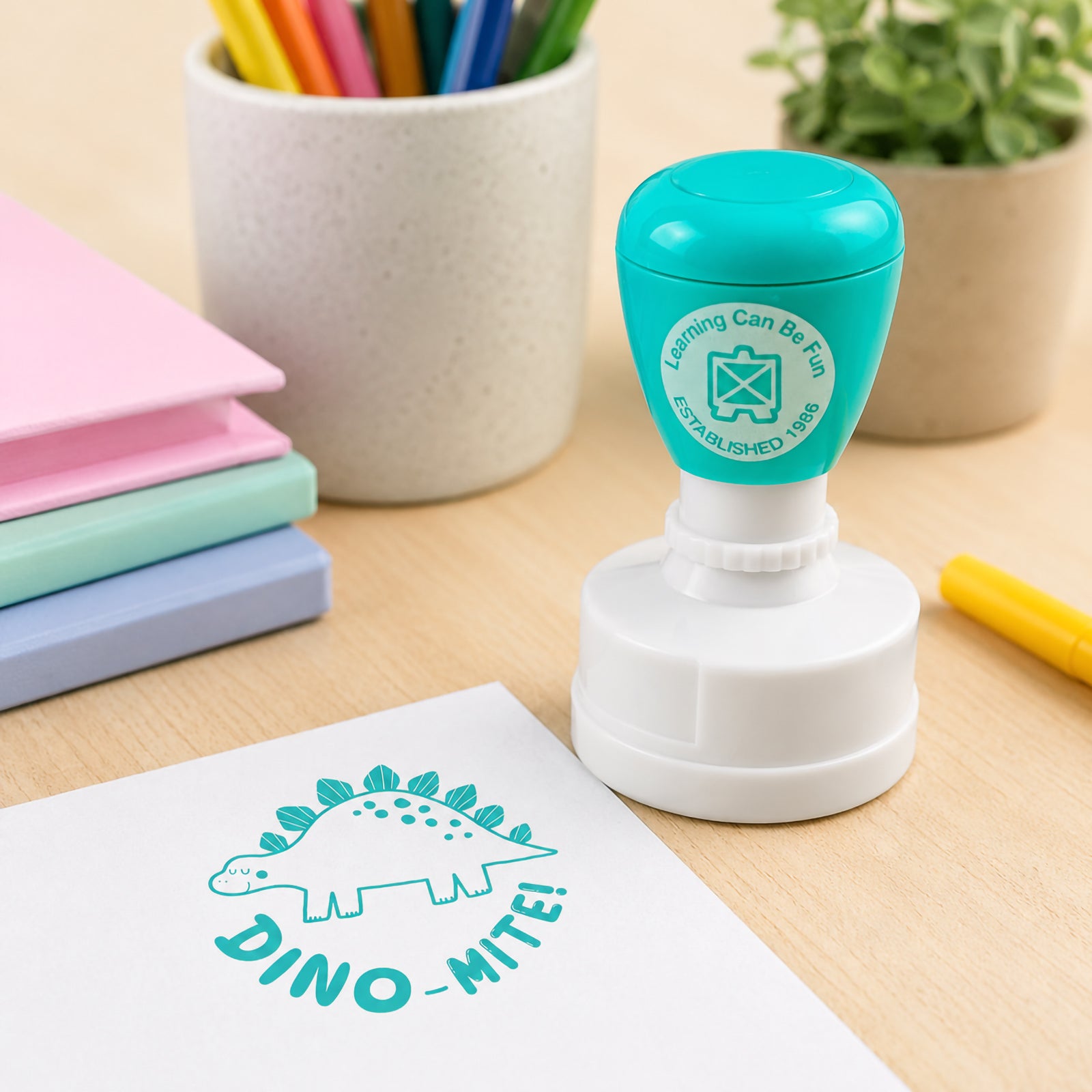 Learning Can Be Fun Dino Mite self inking merit stamp displayed beside colourful books and stationery. The teal dinosaur themed impression appears clearly on paper, creating fun and encouraging classroom feedback for young learners.