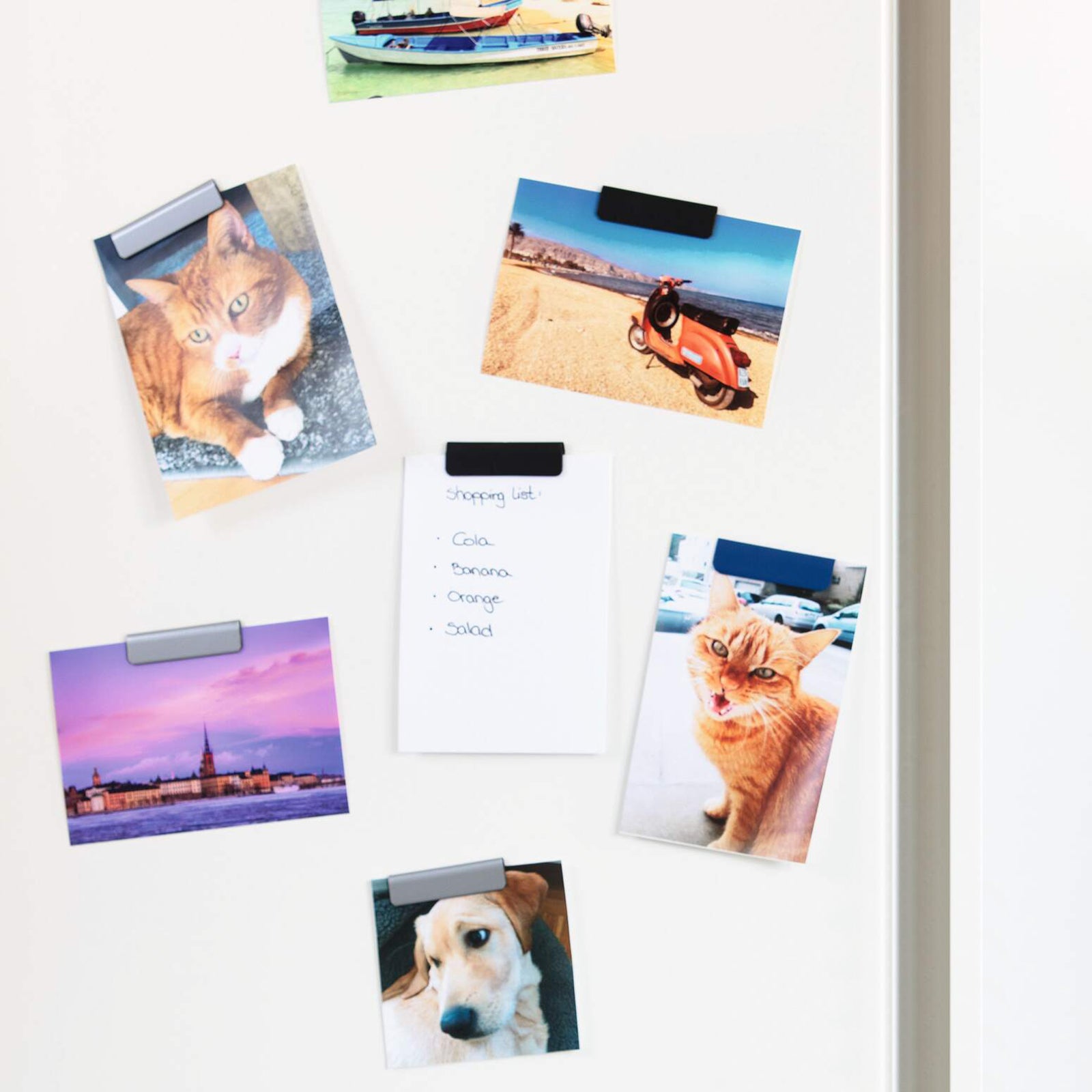 Several Durable Durafix Magnetic Clips in dark blue hold family photographs, postcards and a handwritten shopping list on a white refrigerator door. The clips create a neat, modern display while keeping everyday reminders and treasured memories organised in the kitchen.