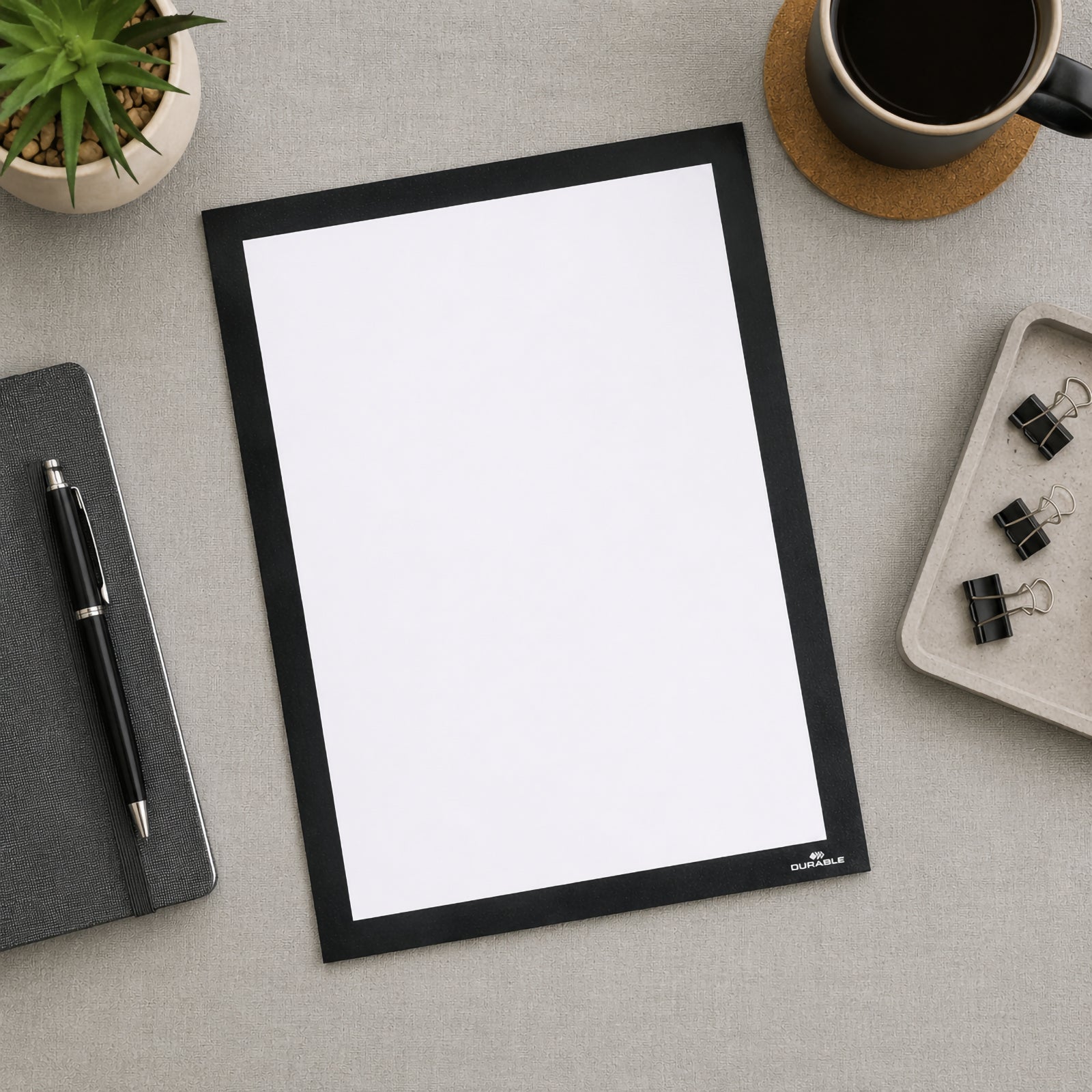 Top view of the Durable DURAFRAME Grip Textile Surface A4 Black placed on a modern office desk beside a notebook, coffee cup, stationery and a potted plant. The blank A4 display frame showcases its premium black finish and professional appearance.