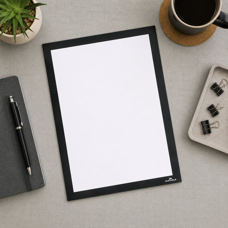 Top view of the Durable DURAFRAME Grip Textile Surface A4 Black placed on a modern office desk beside a notebook, coffee cup, stationery and a potted plant. The blank A4 display frame showcases its premium black finish and professional appearance.