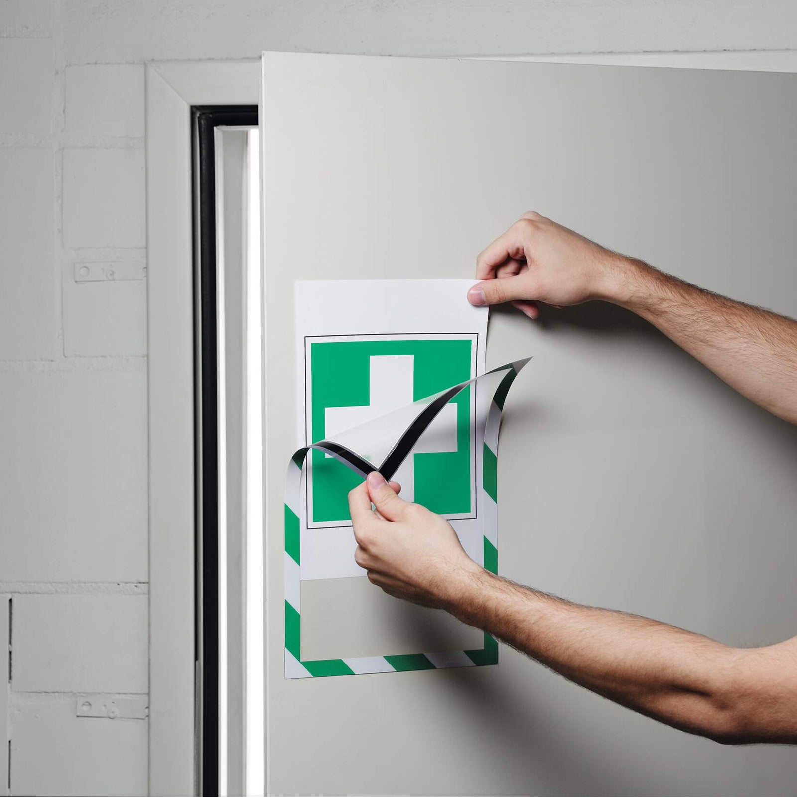 Hands demonstrate replacing an emergency first aid notice inside the Durable DURAFRAME Magnetic Security A4 Green White frame mounted on a metal cabinet. The magnetic front lifts easily for fast document updates without removing the frame from the surface.