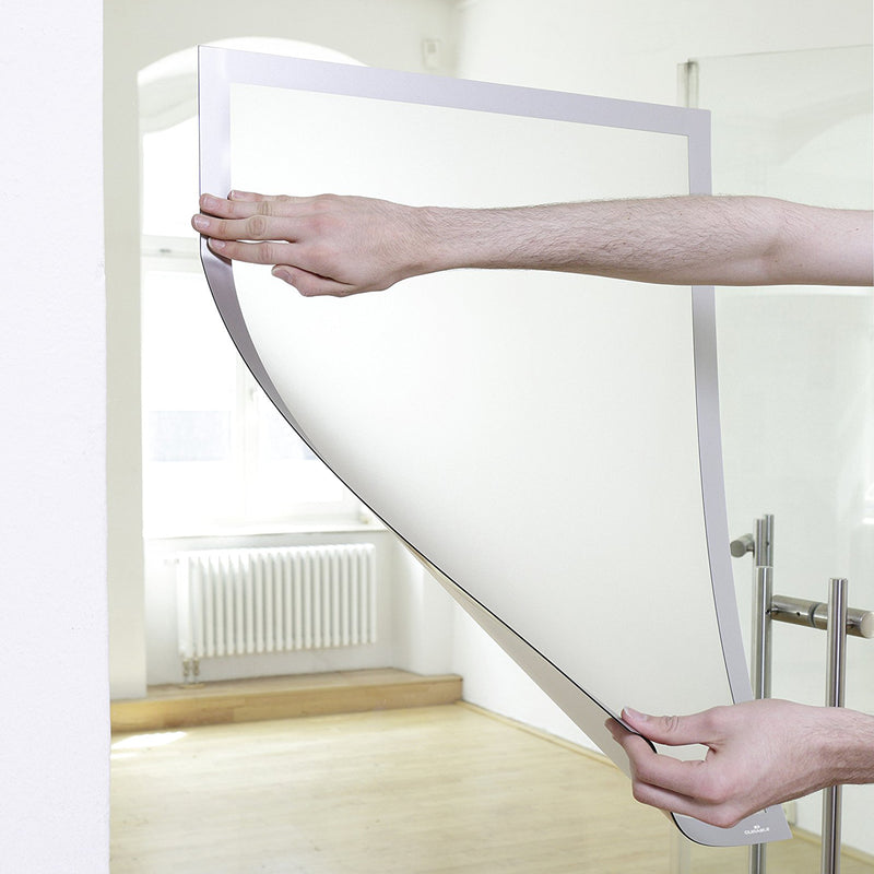 A user applies the Durable DURAFRAME Poster 50 x 70cm Silver to a smooth glass wall by positioning the flexible self adhesive frame. The image demonstrates the easy installation process and large format display area in a bright commercial interior.