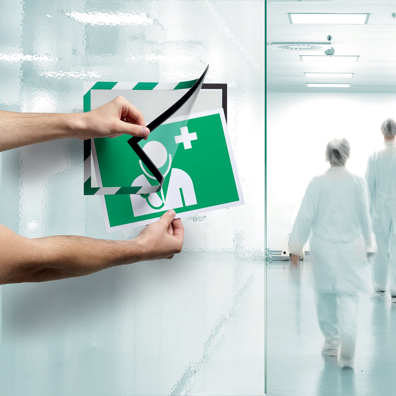 Hands opening the magnetic front of the Durable DURAFRAME Security A4 Green White frame while inserting a first aid information sign onto textured glass. The image demonstrates quick document replacement without removing the frame.
