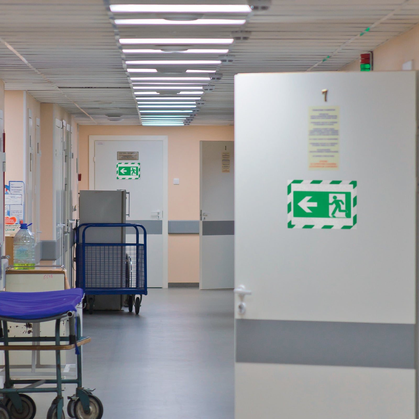 Durable DURAFRAME Security A4 Green White displayed on a hospital corridor door showing an emergency exit direction sign. The highly visible ISO compliant frame provides clear emergency wayfinding in healthcare and public facilities.